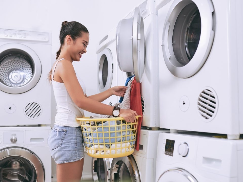 College woman standing and holding a laundry basket. She's putting clothes into a washer