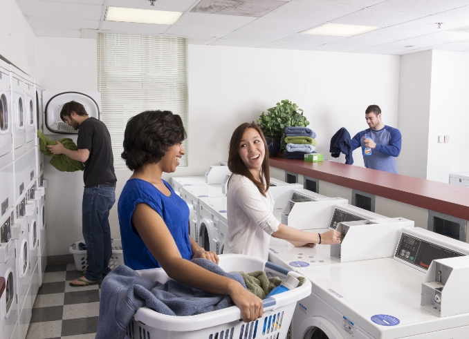 Group of people doing laundry in a community laundry room. 2 men doing laundry on their own and 2 women talking to each other