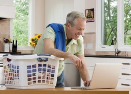 Man working on a laptop with a laundry basket next to him and a shirt over his shoulder