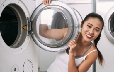 Woman college student in white tank top opening a washer door and smiling at a camera