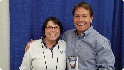 Man and a woman posing together for a photo at a trade show. They're smiling at the camera